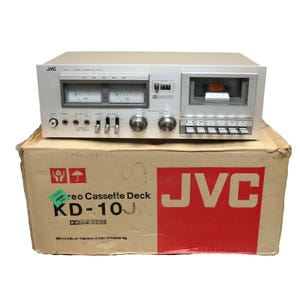 May include: A silver JVC KD-10J stereo cassette deck featuring dual analog VU meters, a cassette compartment, multiple control knobs, and push-button switches. The device sits atop its original cardboard box, which displays the JVC logo and model name.