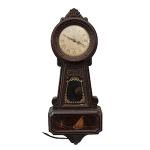 Peut inclure: Une horloge murale en bois brun foncé avec un cadran blanc et des chiffres romains noirs. L'horloge a un design décoratif avec un voilier doré en bas.