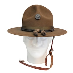 May include: A brown felt hat with a wide brim, a decorative band, and a small emblem. The hat features a leather chin strap and acorn-shaped ornaments. The hat is displayed on a white mannequin head.