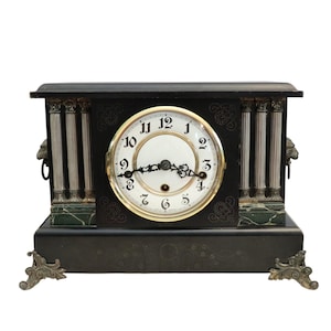 May include: A black mantel clock with a white clock face and gold trim. The clock features silver columns, decorative accents, and ornate feet. The clock's face displays the numbers 1 through 12.