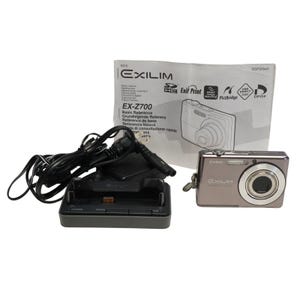 May include: A Casio Exilim EX-Z700 digital camera in silver with a charging dock and instruction manual. The camera has a lens and the word EXILIM is printed on the front. The instruction manual has the camera's model number and features.