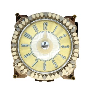 May include: A circular clock face with a pale yellow center and a silver-toned rim. The dial features dark blue Arabic numerals from 1 to 12. A decorative border surrounds the dial, consisting of alternating cream-colored and silver-toned segments.