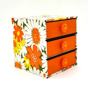 May include: A small orange and white floral patterned dresser with three drawers and orange knobs.