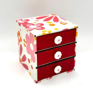 May include: A small, three-drawer jewelry box. The box is white with a floral pattern in pink, orange, and yellow. The drawers are red with white flower-shaped knobs. The box is a decorative storage item.