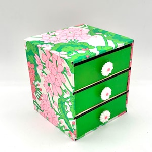 May include: A small green three-drawer box with white floral print on the front and white flower knobs on each drawer.