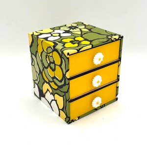 May include: A small, three-drawer dresser with a floral pattern in shades of green, yellow, and white. The drawers are yellow with white daisy-shaped knobs.