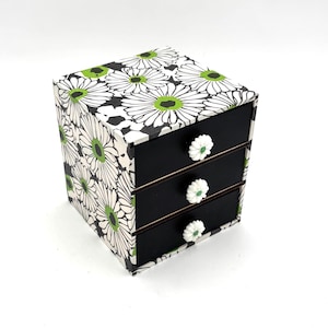 May include: A small square storage chest with three stacked drawers. The exterior features a black and white daisy floral pattern with bright green centres. Each drawer has a solid black front and a white flower-shaped knob for opening.
