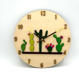 May include: A wooden clock with a cactus design. The clock face is light brown with gold numbers and hands. The cactus are green, yellow, and pink.