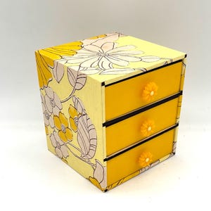 May include: A small yellow wooden box with three drawers. The box is decorated with a floral pattern in shades of yellow and white. The drawers have yellow knobs.