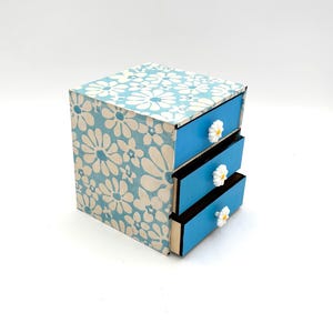 May include: A miniature three-drawer chest. The box features a blue and white floral design on the top and sides. The drawers are a solid blue with white daisy-shaped knobs. The box is likely made of wood or a similar material.