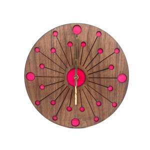 May include: A round wooden clock with a pink and gold design. The clock face has a sunburst pattern with pink circles around the center. The clock hands are gold.