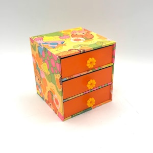 Peut inclure: Petite boîte à trois tiroirs avec un extérieur à motifs colorés. La boîte a un fond jaune avec des personnages de dessins animés et des tiroirs orange avec des boutons en forme de fleur jaunes. La boîte est probablement destinée à ranger de petits objets.