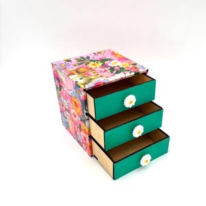 May include: A small wooden drawer organiser with three drawers. The drawers are painted teal green and have white daisy-shaped knobs. The exterior of the organiser is covered in a floral print with pink, yellow, and green flowers.