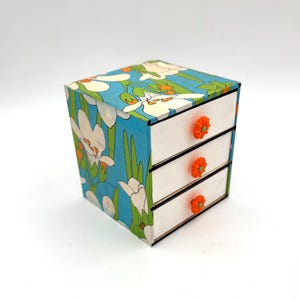 May include: A small white wooden chest of drawers with three drawers. The drawers have orange flower-shaped knobs. The chest of drawers is covered in blue and green floral patterned paper.
