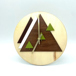 May include: A round wooden clock with a light wood grain background. The clock face features a dark brown triangle, a white triangle, and green tree shapes. The clock hands are gold.