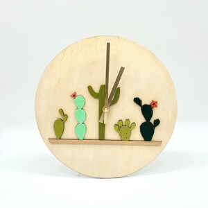 May include: A wooden clock with a cactus design. The clock face is made of light wood and features a gold-colored hour and minute hand. The cactus design is made of green, red, and brown felt and is arranged in a row across the bottom of the clock face.