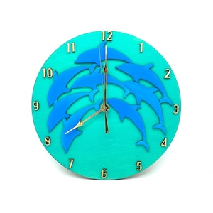 Puede incluir: Un reloj redondo azul turquesa con un diseño de delfines azules saltando en círculo. La esfera del reloj tiene números dorados del 1 al 12 y manecillas doradas.
