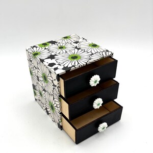 May include: A small white and black floral patterned wooden box with three drawers. Each drawer has a white knob with a green center.