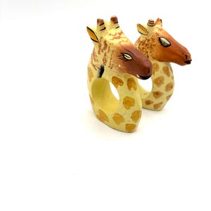 May include: Two wooden giraffe napkin rings. The giraffes are painted yellow with brown spots and have brown faces.