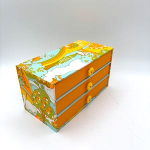 May include: A small, three-drawer chest with a vibrant, retro floral pattern on the exterior. The drawers are orange with light blue trim and yellow flower-shaped knobs. The chest is a decorative storage item.