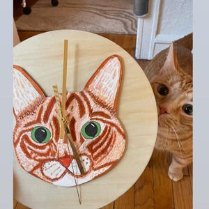 May include: A wooden clock featuring a hand-painted orange tabby cat face. The cat has green eyes and a pink nose. A real orange tabby cat is looking at the clock.