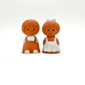 May include: Two terracotta figurines of children. One wears orange overalls with a white shirt, the other a white dress with a bow. Both have round heads, black eyes, and are set against a white background.