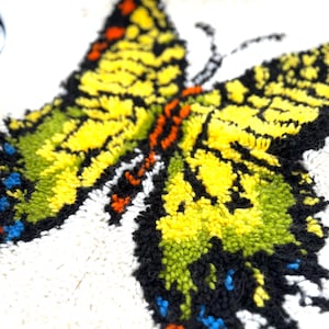 May include: Close-up of a colorful butterfly design. The butterfly features yellow, green, orange, black, and blue accents. The design is made with a textured, looped material, set against a white background.