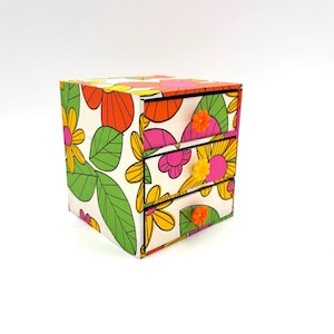 May include: A small three-drawer storage chest featuring a white background with a vibrant floral pattern in orange, yellow, pink, and green. Each drawer is equipped with a small, flower-shaped knob in either orange or yellow plastic.