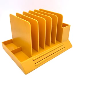 May include: A yellow plastic desk organiser with multiple vertical dividers for files, a small compartment, and a pen holder. The organiser is rectangular and has a simple, functional design.