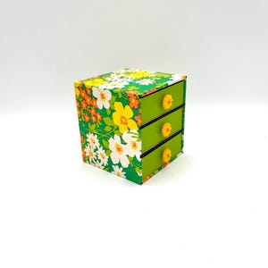 May include: A small green three-drawer dresser with floral print and yellow and orange flower accents. The drawers have yellow plastic knobs.