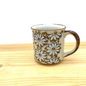 May include: A ceramic mug with a speckled off-white glaze featuring a repeating pattern of blue-outlined daisy flowers and brown accents. The mug has a cylindrical body, a smooth rim, and a curved handle with a dark brown glazed finish.