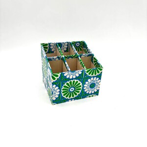 May include: A green, blue, and white patterned cardboard organizer with six compartments. The organizer is perfect for storing small items like pens, pencils, or makeup brushes.