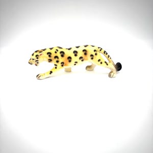 May include: A small ceramic figurine of a leopard in a prowling stance. The body is cream-colored with dark brown and black spots distributed across its back and sides. The leopard has a long tail with a black tip and a detailed facial expression.