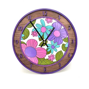 May include: A round wall clock with a wooden frame and a purple border. The clock face features a floral pattern in pink, purple, and turquoise. The clock hands are black.