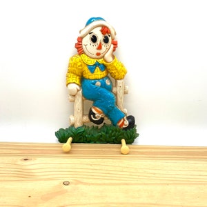 May include: A vintage scarecrow figurine with a yellow shirt, blue pants, and a red hat. The scarecrow is sitting on a wooden fence with green bushes at the bottom. The figurine has two wooden hooks for hanging items.