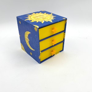 May include: A small, three-drawer jewelry box with a celestial theme. The box is blue with illustrations of the sun, moon, and stars. The drawers are yellow with star-shaped knobs. The box is approximately 4 inches wide, 4 inches deep, and 4 inches tall.