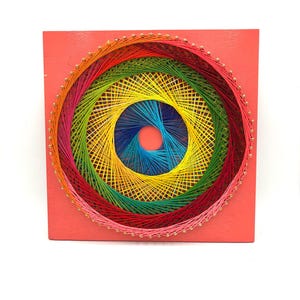 May include: A square string art piece on a coral-colored wooden board featuring a circular pattern created with colorful threads. The design uses concentric rings of pink, red, green, yellow, and blue thread wrapped around small metal nails.