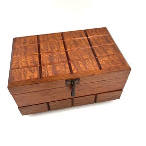 May include: A rectangular wooden box with a rich brown grain, featuring a grid-like pattern of carved squares on the lid and lower section. The box includes a dark metal latch centered on the front, which secures the lid to the main body.