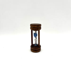 May include: A small hourglass with a wooden frame and base. The hourglass contains blue sand and is set against a white background. The wooden frame has a dark brown finish.
