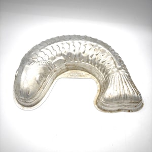 Puede incluir: Un molde para pastel vintage en forma de pez, de color plateado. El molde tiene escamas, aletas y una cola detalladas. El molde es de metal y tiene un asa. La marca "Mirro" es visible.