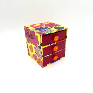 May include: A small, yellow, floral patterned drawer chest with three drawers. Each drawer has a red front and a bright orange flower-shaped knob.