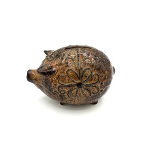 May include: A brown, textured ceramic piggy bank featuring a dark, weathered finish and a prominent, etched floral pattern on its side. The pig-shaped coin bank has a slot on its back for inserting coins and displays a rounded, compact form.