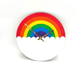 May include: A circular wall clock featuring a colorful rainbow design with concentric bands of red, orange, yellow, green, blue, and purple. The bottom portion of the clock is a solid white cloud shape. It has two thin gold-colored metal hands.