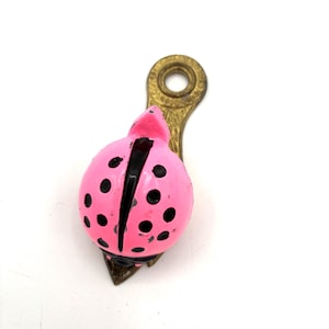 May include: A pink ladybug-shaped decorative clip with black spots and a brass-colored metal attachment. The ladybug has a black line down the center. The clip is on a white background.