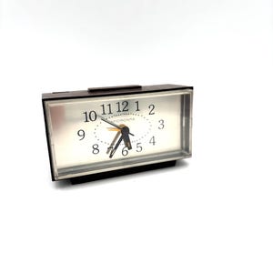 May include: A rectangular analog alarm clock with a dark brown frame and a clear plastic front cover. The clock face is silver with black Arabic numerals from 1 to 12, black hour and minute hands, a thin orange second hand, and the text MICRONTA.