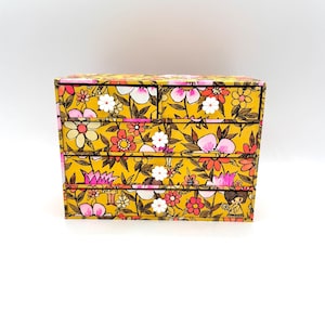 May include: A small, decorative chest of drawers with a vibrant floral pattern in yellow, pink, white, and brown. The chest has four drawers and a top compartment, perfect for storing small items. The design features a repeating pattern of stylized flowers and leaves.