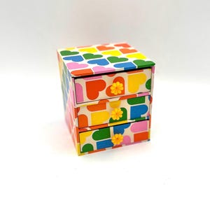 May include: A small, colourful dresser with three drawers. The drawers have yellow flower-shaped knobs and are decorated with a pattern of multi-coloured hearts.