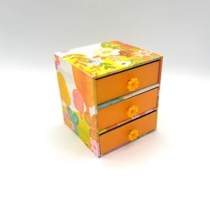 Peut inclure: Petite boîte à trois tiroirs, orange à l'extérieur, avec un motif floral sur le dessus et les côtés. Les tiroirs sont orange avec des boutons en forme de fleur jaune. Objet de rangement décoratif.