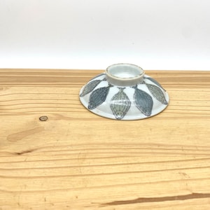 May include: A small white ceramic bowl with a blue and green fish design. The bowl is sitting on a wooden surface.