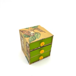 May include: A small, square, three-drawer jewellery box. The box has a light brown background with owl illustrations and green drawers. Each drawer has a yellow flower-shaped knob. The box is on a white background.